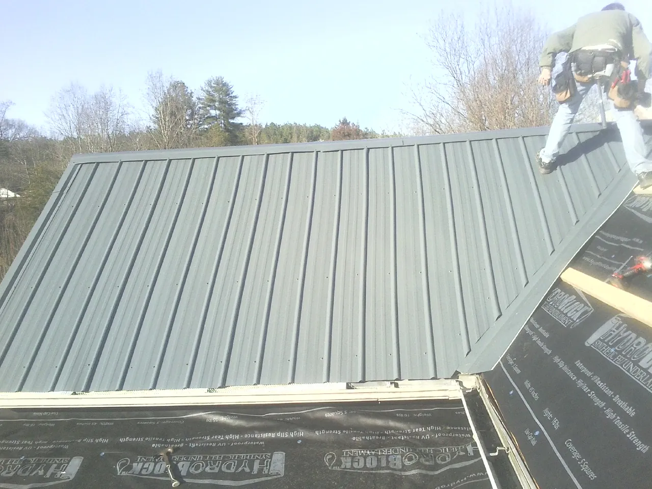 Crew installing dark green standing seam panels over synthetic underlayment in Clarion