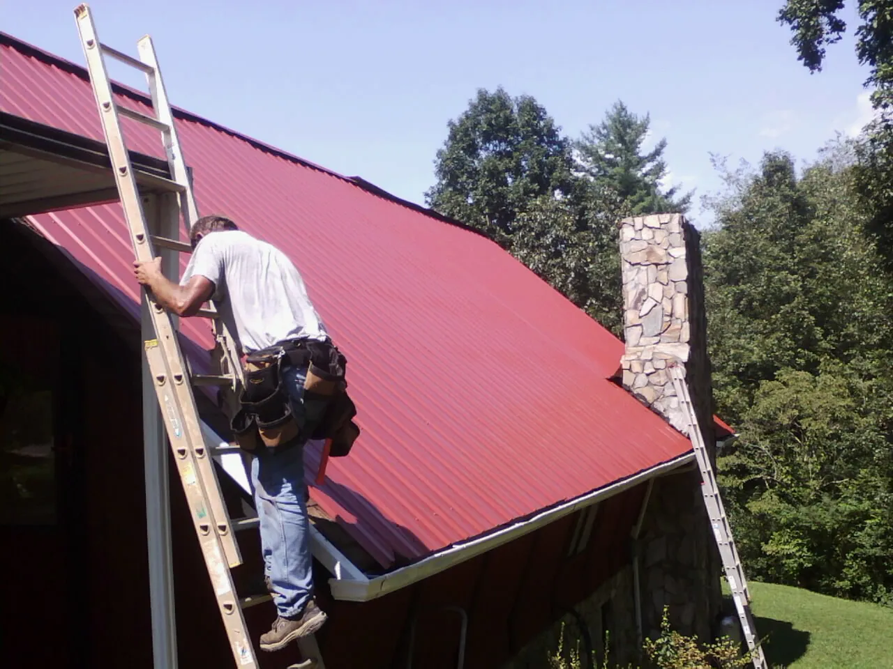 Roofer on a ladder beside a red metal roof and stone chimney — Soffit & Fascia Repair by Roofing Brothers in Clarion
